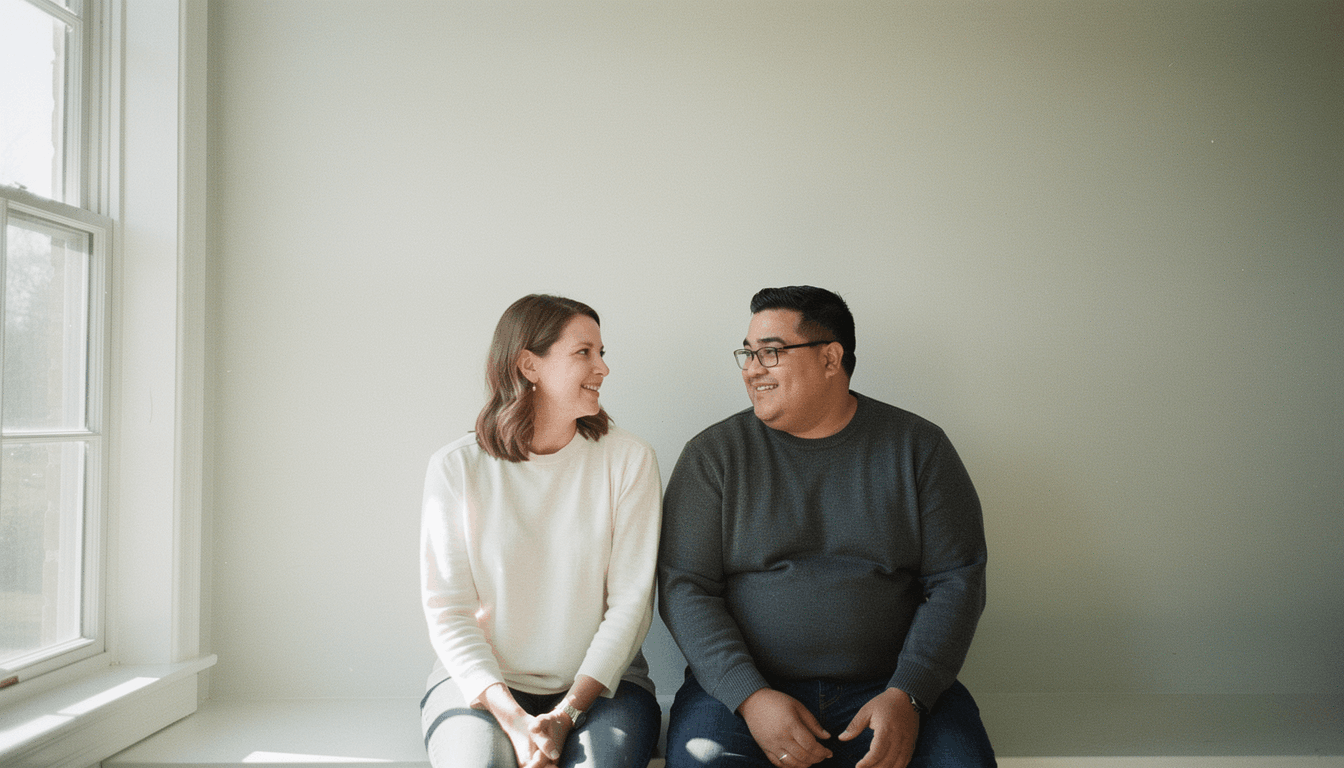 A couple sits together on a bright windowsill, talking, one holding a purple mug.