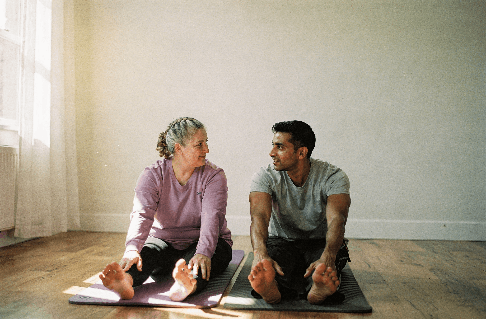 Two friends sit on exercise mats stretching in a sunlit living room.