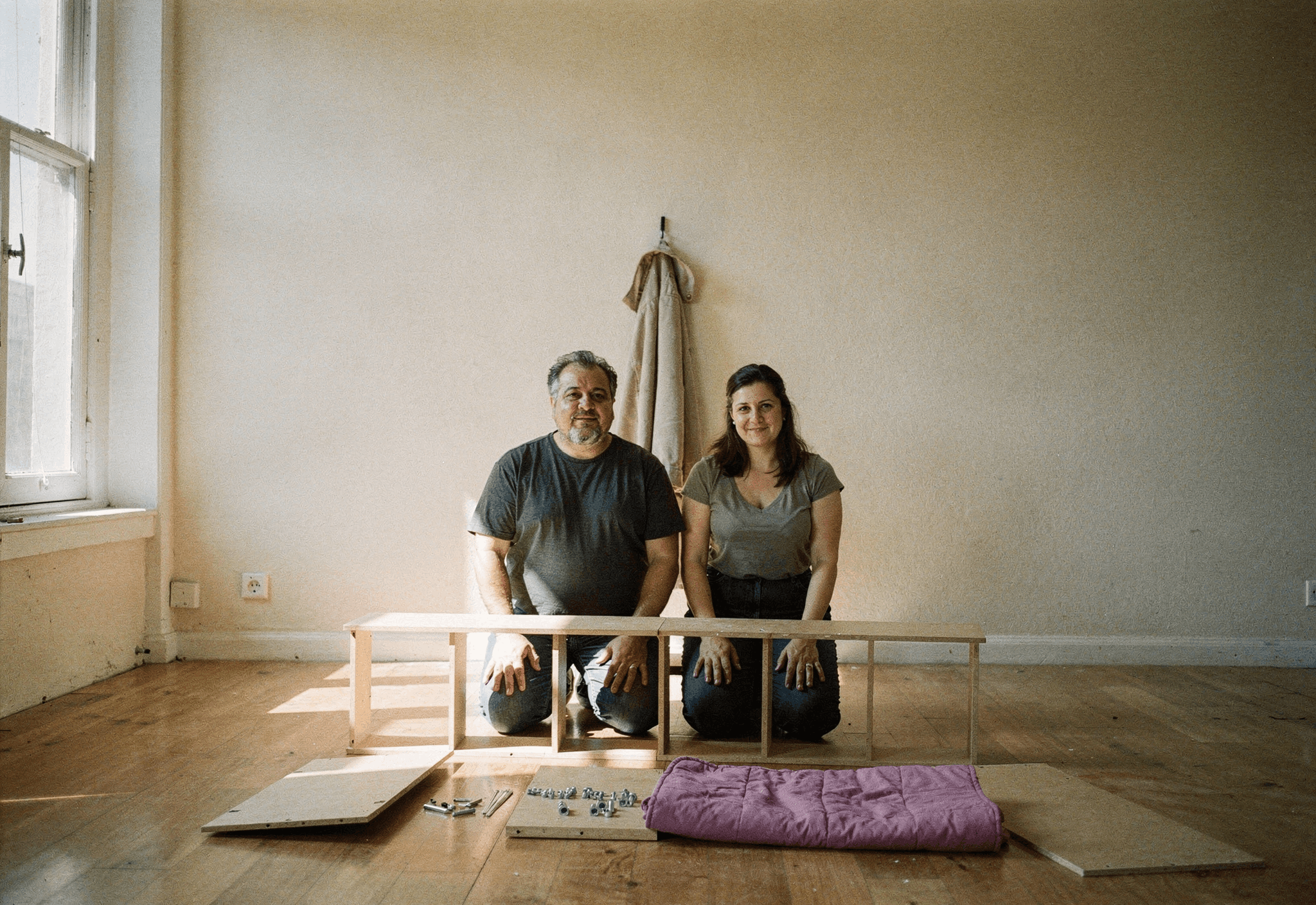 Two neighbors kneel on a bare floor assembling a flat-pack bookcase in a sunlit empty apartment.
