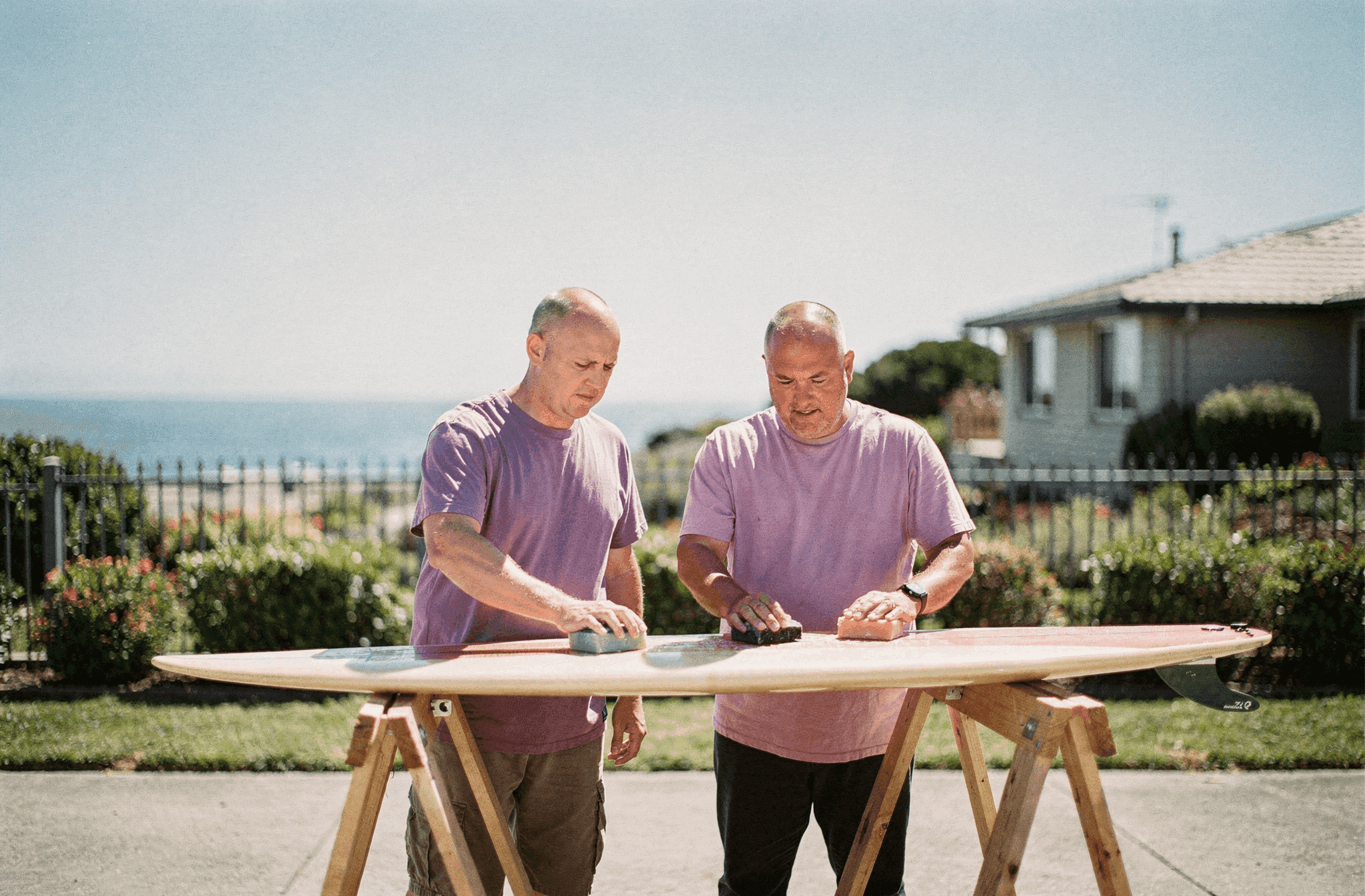 Two neighbors wax a surfboard together on sawhorses on a bright day.