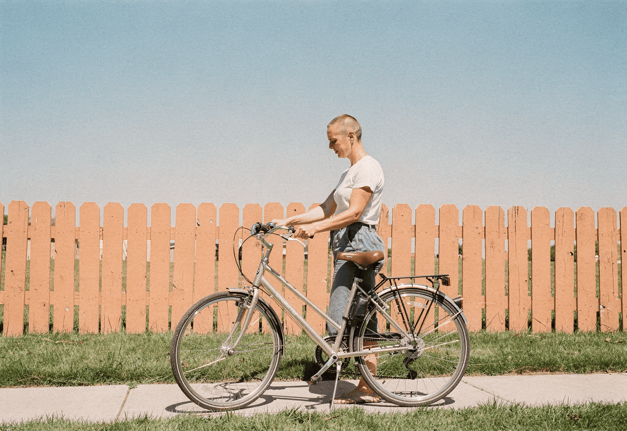 A woman in her late thirties fits a bell onto her bike's handlebars beside an orange picket fence.