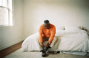 A man in his late fifties sits on the edge of a bed putting on a walking shoe in a bright bedroom.