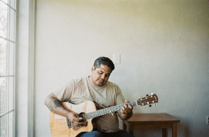 A man in his late forties restrings an acoustic guitar in a bright room.