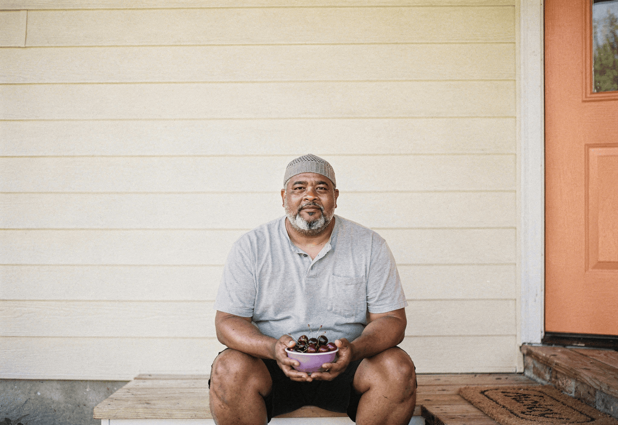 A man in his early sixties sits on his front porch step with a bowl of cherries in late-afternoon sun.