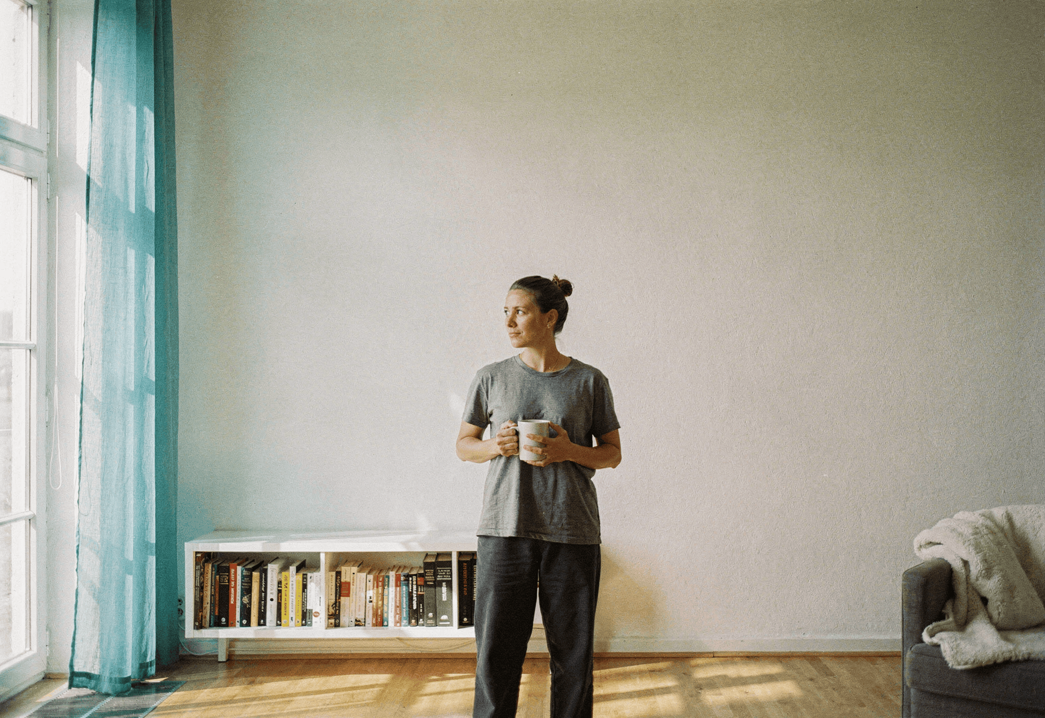A person in their late forties holds a mug beside a sunny window with a sheer teal curtain.