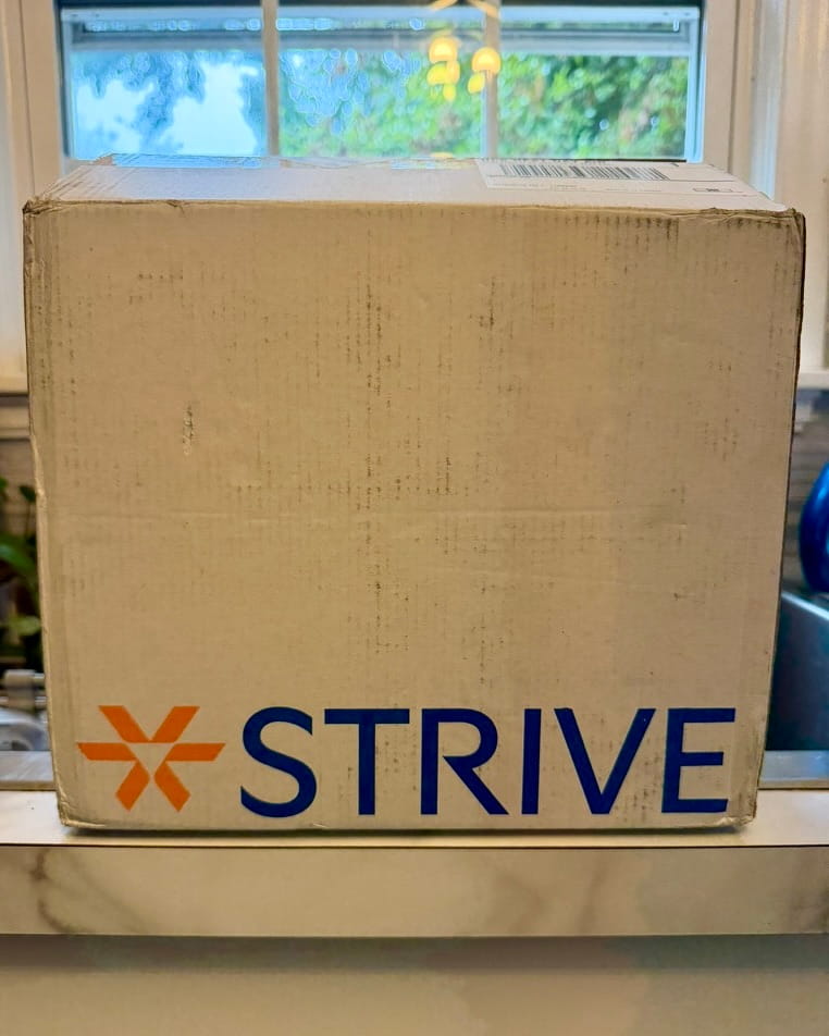 Plain kraft Strive Pharmacy shipping box with an orange asterisk logo and the word STRIVE standing on a kitchen counter in front of a window