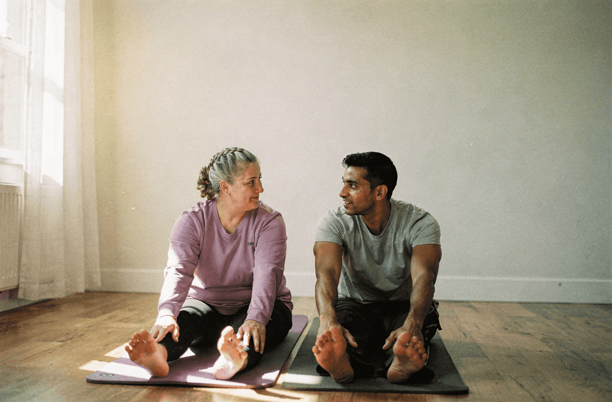 Two friends sit on exercise mats stretching in a sunlit living room.