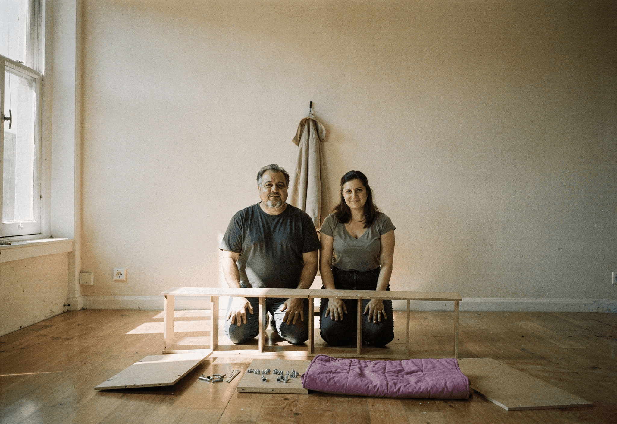 Two neighbors kneel on a bare floor assembling a flat-pack bookcase in a sunlit empty apartment.