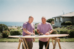 Two neighbors wax a surfboard together on sawhorses on a bright day.