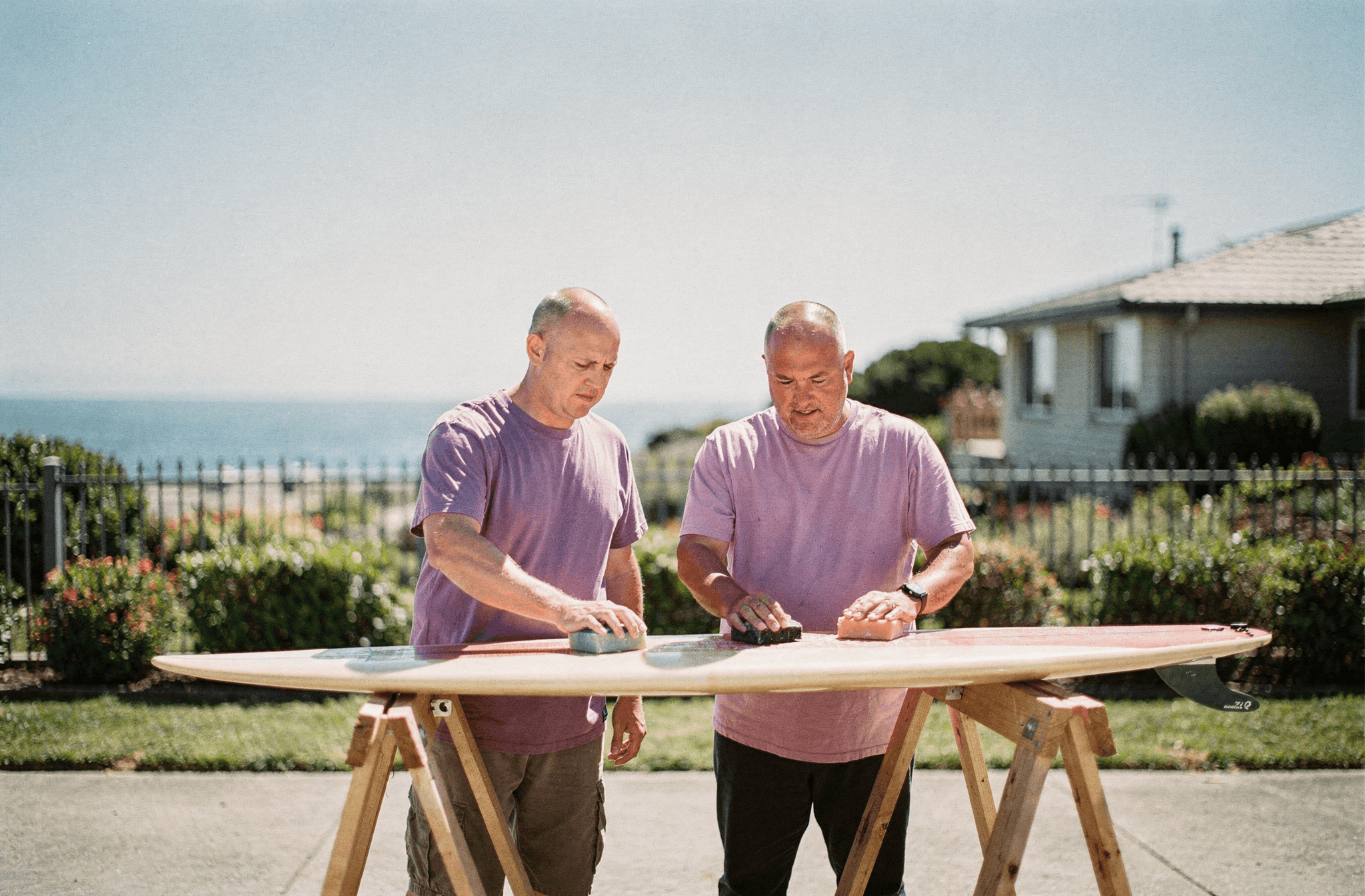 Two neighbors wax a surfboard together on sawhorses on a bright day.
