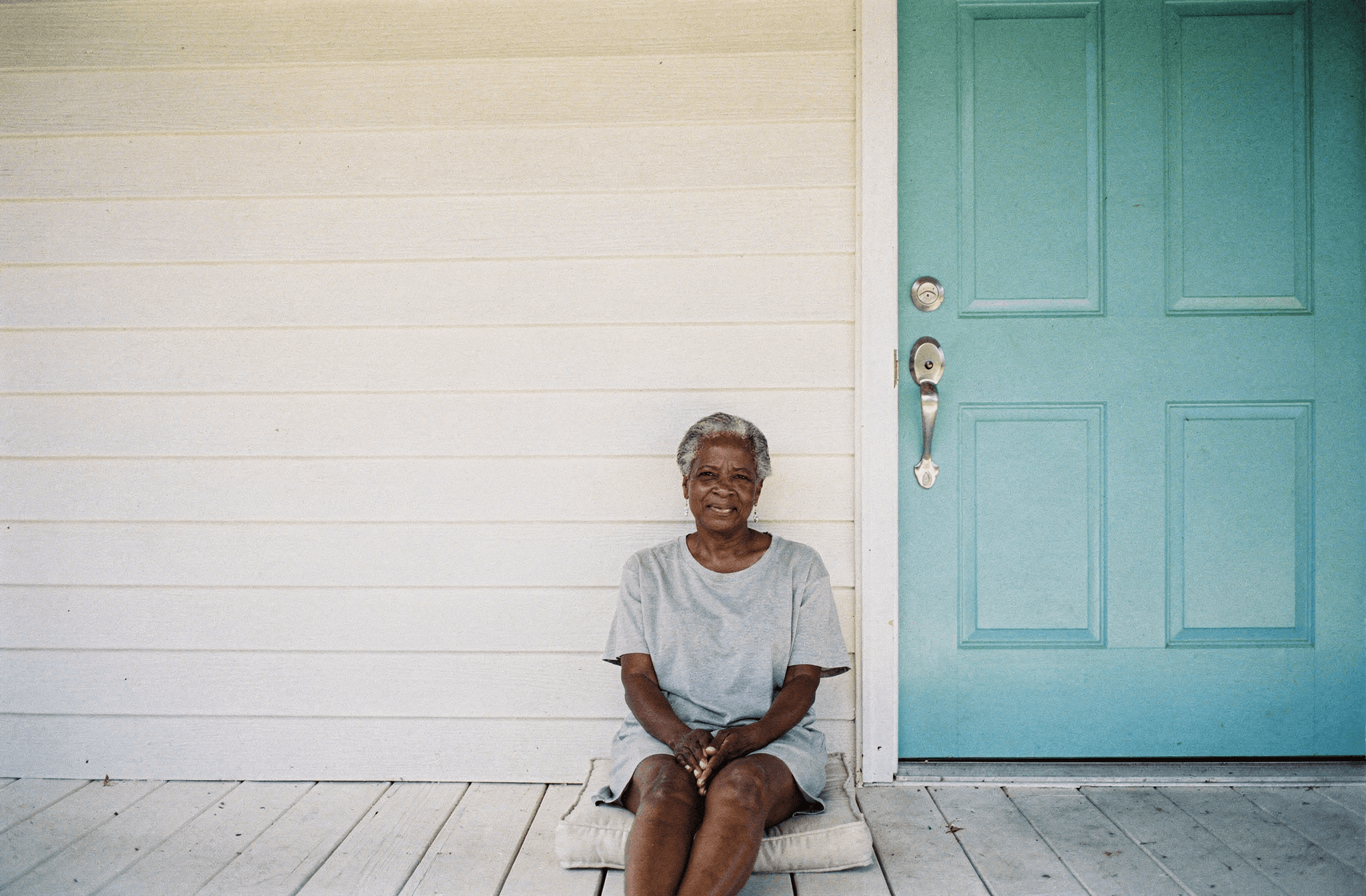 A woman in her late fifties sits on her front step beside a teal painted door.