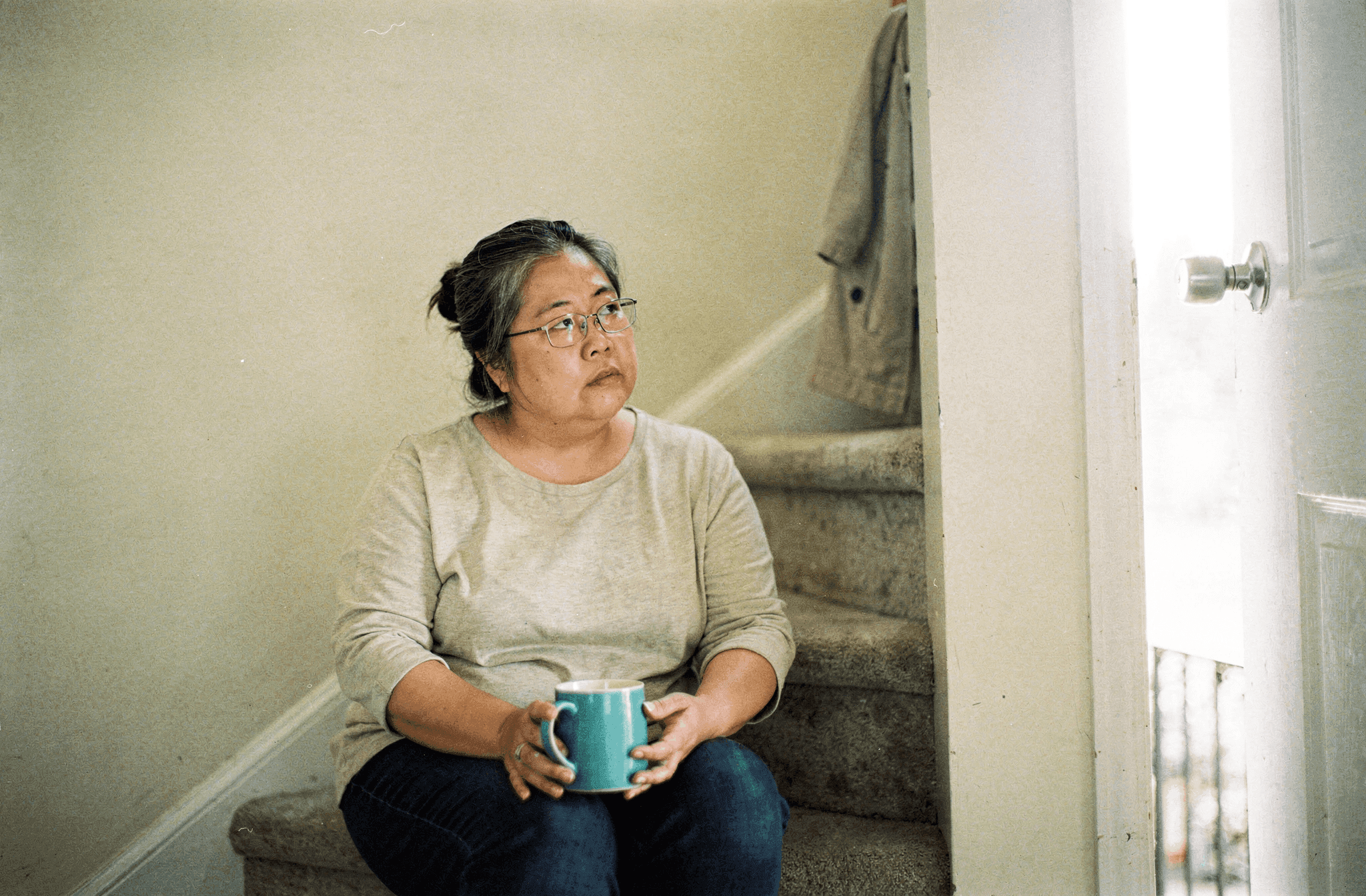 A woman in her late forties sits on a stair landing holding an aqua mug in both hands.